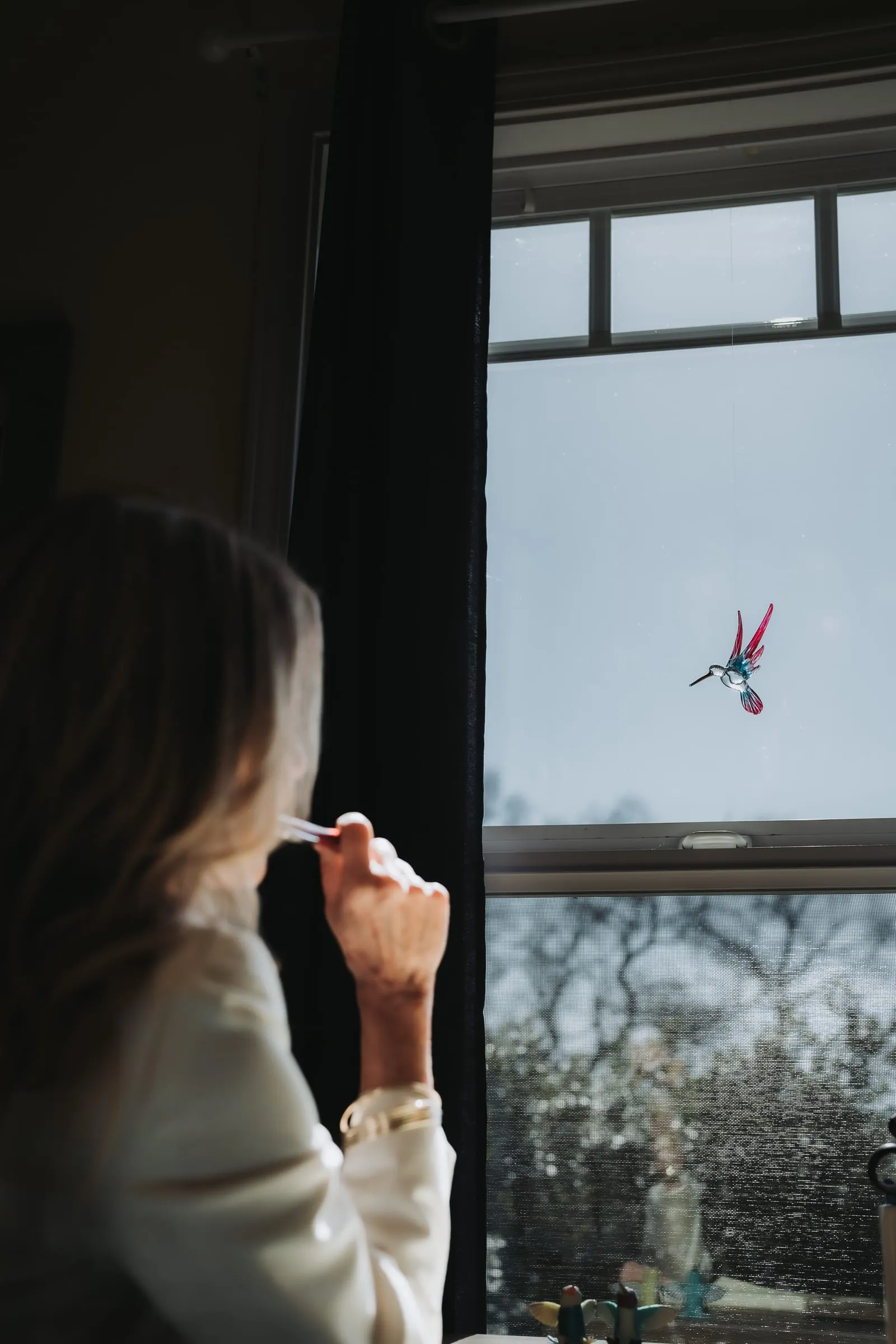 Woman pausing in thought at a bright window, a glass hummingbird hanging from the frame