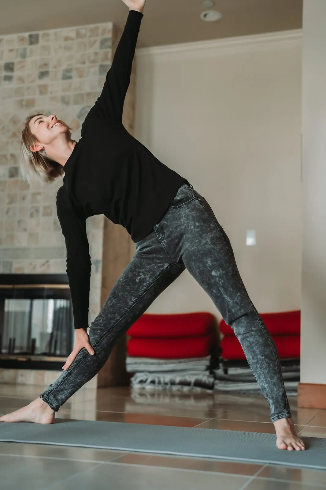 A woman stretches into a standing triangle pose on a yoga mat beside a stone fireplace